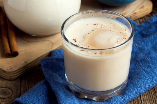 Milky white drink with ice and a sprinkling of cinnamon on a blue towel with cinnamon sticks and a pitcher of milk in background